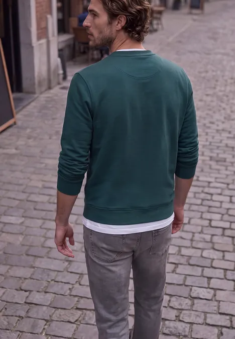 Ein Mann von hinten in einem grünen Pullover und grauen Hosen auf einer Kopfsteinpflasterstraße.