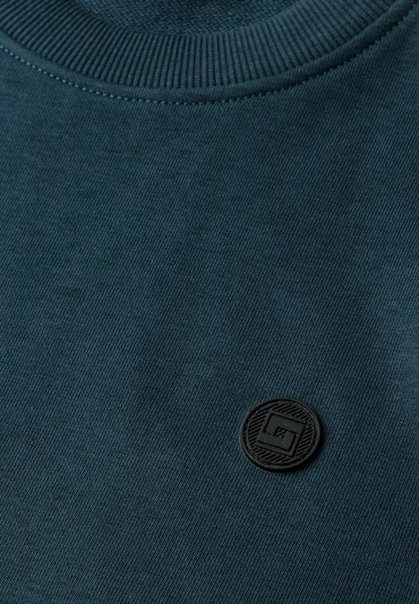 Dunkelgrünes Shirt mit rundem, schwarzem Logo-Aufnäher an der Brust.