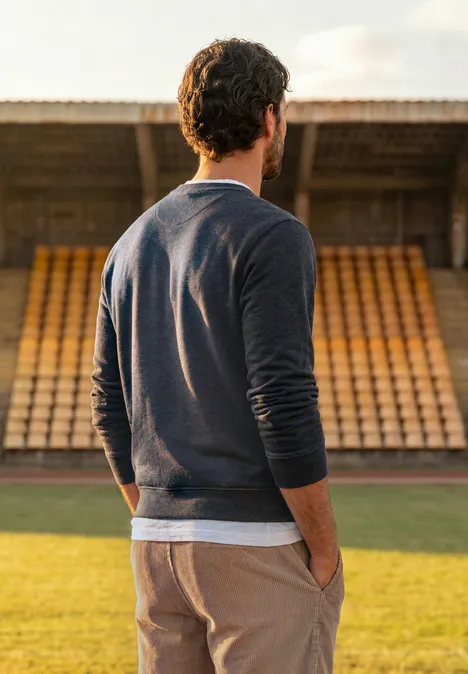Ein Mann im Pullover steht auf einem Stadionrasen und blickt auf leere Tribünen.