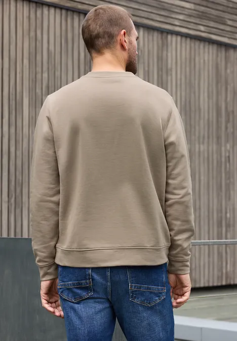 Ein Mann von hinten in graubraunem Pullover vor holzvertäfelter Fassade.