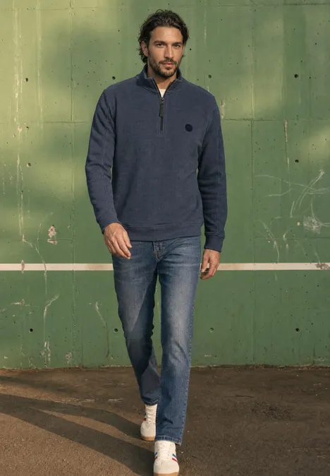 Sweatshirt à col montant et fermeture zip dark ink blue Ein Mann in Jeans, weißen Turnschuhen und blauem Zip-Pullover geht vor einer grünen Wand mit weißer Linie.