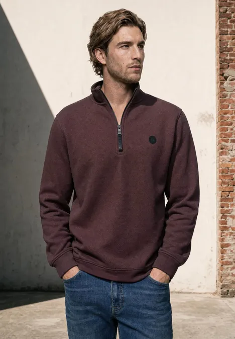 Sweatshirt à col montant et fermeture zip maroon brown Ein Mann trägt einen bordeauxroten Pullover mit halbem Reißverschluss und steht vor einer Backsteinwand.