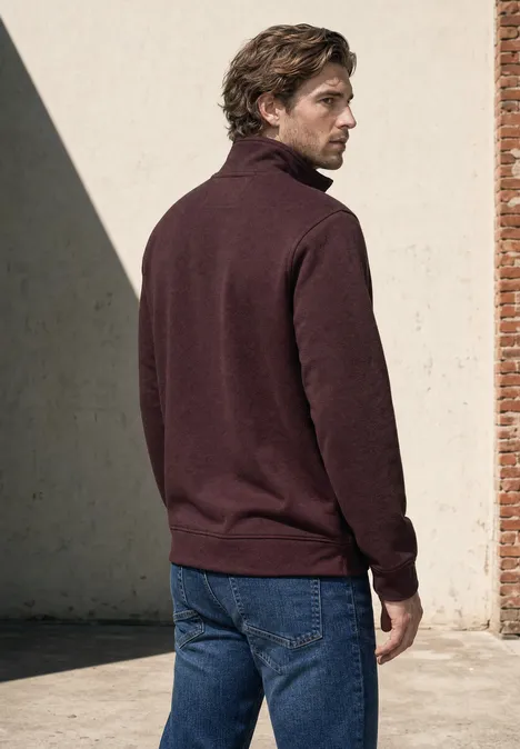 Sweatshirt à col montant et fermeture zip maroon brown Mann mit braunem Rollkragenpullover und Jeans steht seitlich vor einer Wand mit einem Ziegelpfeiler.