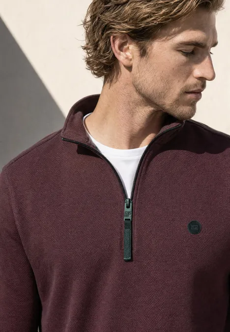 Sweatshirt à col montant et fermeture zip maroon brown Ein Mann in einem burgunderroten Pullover mit Reißverschluss und kleinem Logo, der nach unten blickt.