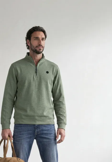 Ein Mann mit Bart trägt einen olivgrünen Pullover und Jeans, steht vor hellem Hintergrund und hält zwei Beuteltaschen.