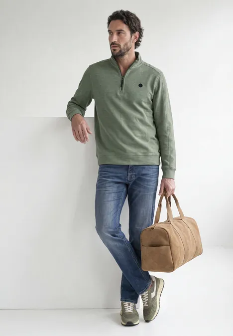 Ein Mann in grünem Pullover, Jeans und Sneakers lehnt an einer weißen Wand, trägt eine beige Reisetasche.