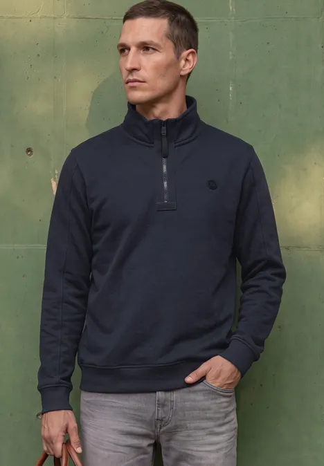 Ein Mann in dunklem Stehkragenpullover mit Reißverschluss steht vor einer grünen Wand und hält etwas in der Hand.
