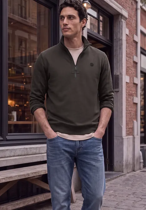Ein Mann steht vor einem Gebäude, trägt ein dunkelgrünes Langarmshirt mit Reißverschluss und Jeans.