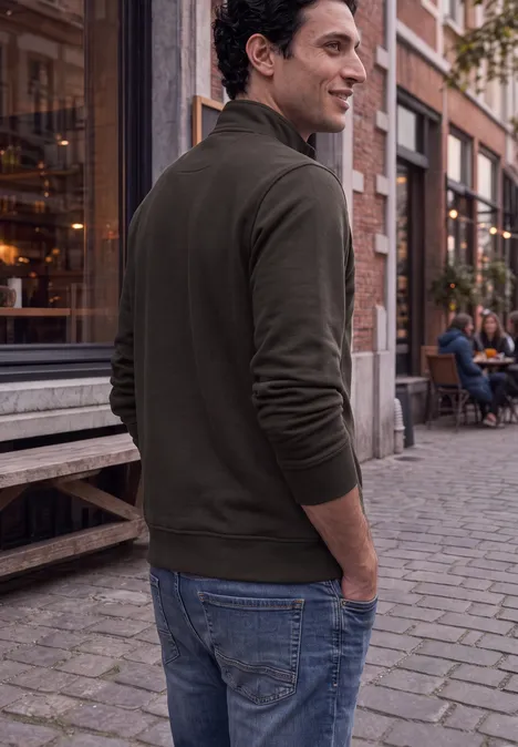 Ein Mann in Jeans und grünem Pullover blickt über die Schulter, steht auf einer Kopfsteinpflasterstraße vor Cafés.