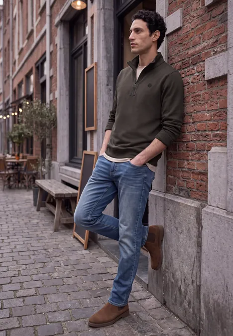 Ein Mann lehnt lässig an einer Backsteinwand in einer Kopfsteinpflastergasse, trägt Pullover und Jeans.