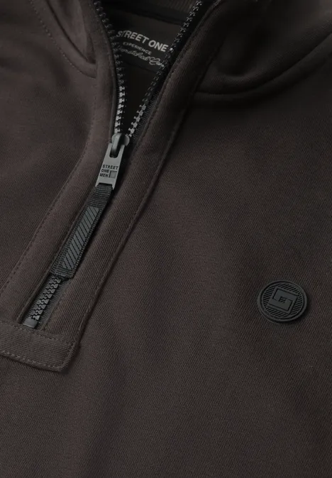 Detailaufnahme einer dunkelbraunen Jacke mit Reißverschluss und rundem Logo auf der Brust.