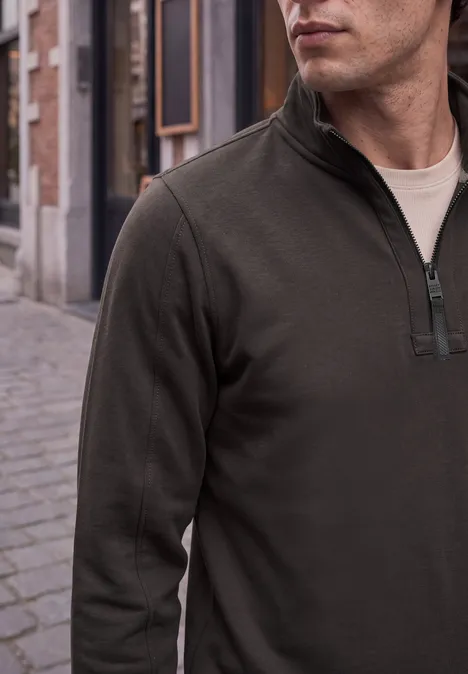Mann in dunkelgrünem Halbreißverschluss-Pullover mit Stehkragen steht auf Kopfsteinpflaster.
