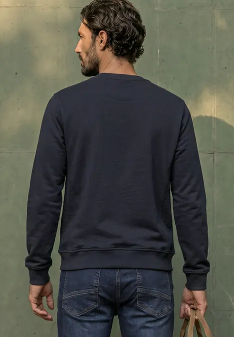 Ein bärtiger Mann im dunkelblauen Pullover und Jeans steht mit dem Rücken zur Kamera, hält eine braune Tasche.