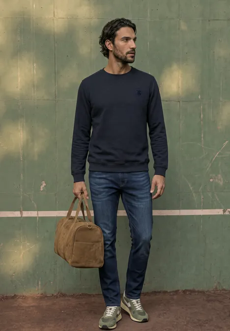 Ein Mann in dunkelblauem Pullover, Jeans und Sneakers steht vor grüner Wand und hält eine braune Tasche.