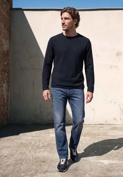 Een man in jeans en donkere trui loopt voor een muur.