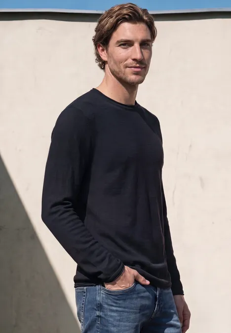 Man in blauw sweatshirt en spijkerbroek voor een lichte muur.