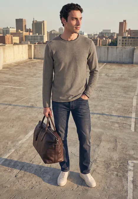 Man in jeans en grijze trui staand op parkeerdak, bruine tas in hand, stedelijke skyline op de achtergrond.