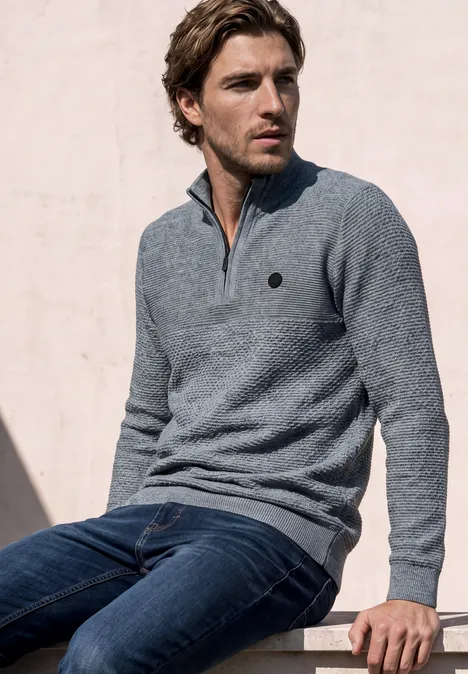 Pullover mit Troyerkragen und Struktur vintage blue Ein junger Mann mit grauem Pullover und Jeans sitzt entspannt auf einer Mauer vor hellem Hintergrund.
