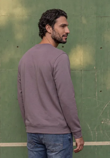 Ein Mann in lila Pullover und Jeans steht vor einer grünen Wand, Rückenansicht, dreht den Kopf seitlich.