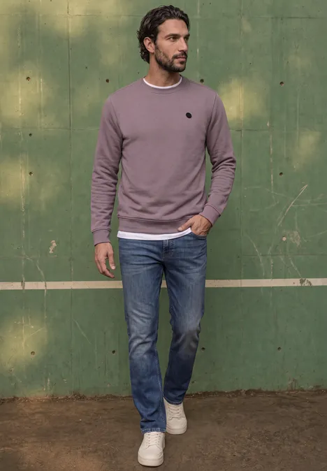 Ein Mann in Jeans, weißem T-Shirt und beerenfarbenem Pullover spaziert vor einer grünen Wand.
