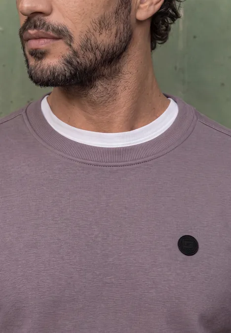 Nahaufnahme eines Mannes mit kurzem Bart, der einen fliederfarbenen Pullover über weißem T-Shirt trägt.