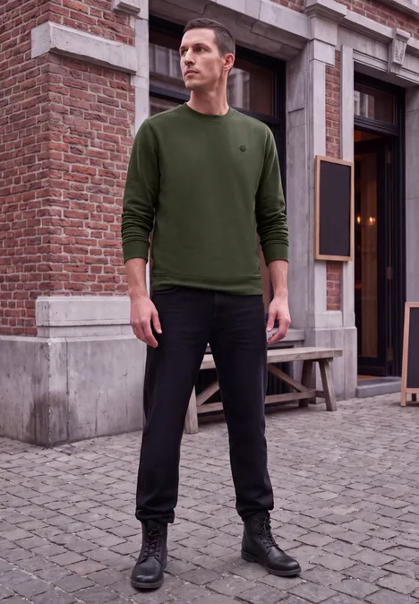 Ein Mann in grünem Pullover, schwarzer Hose und Boots steht auf gepflastertem Gehweg vor einem Backsteingebäude.
