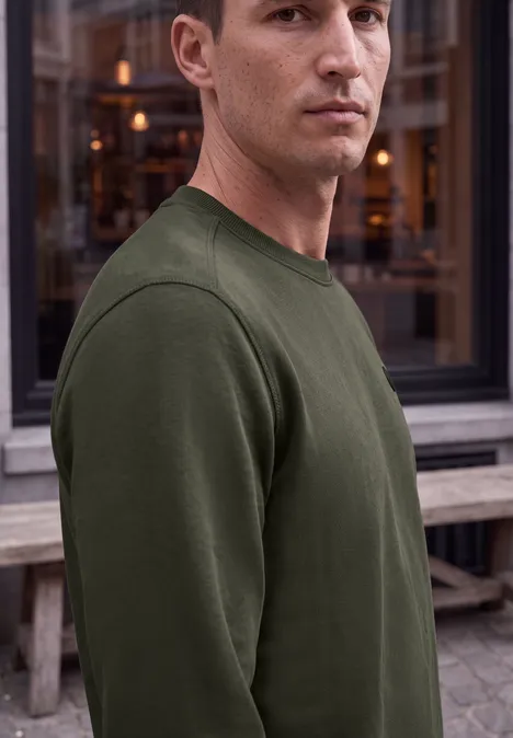Ein Mann im olivgrünen Pullover steht seitlich vor einem Fenster und blickt in die Kamera.