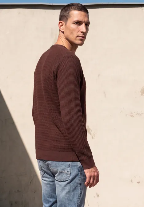 Ein Mann steht vor einer hellen Wand, trägt braunen Pullover und Jeans und blickt über die Schulter.