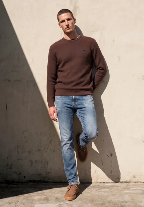 Ein Mann lehnt lässig an einer Wand, trägt braunen Pullover, blaue Jeans und braune Schuhe.