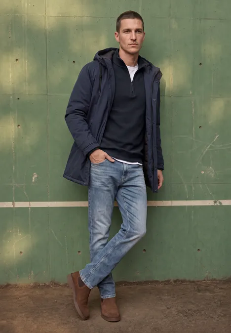 Een man leunt nonchalant tegen een groene muur, gekleed in jeans, bruine laarzen en een donkerblauwe jas.