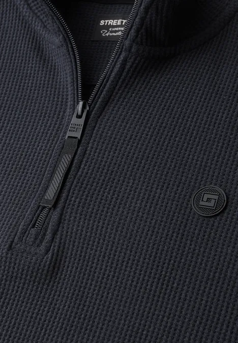 Close-up van een zwart fijngebreid shirt met halve ritssluiting en honingraatstructuur, voorzien van een klein logo.
