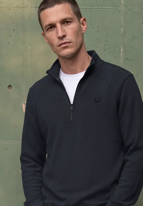 Een man voor een groene metalen wand draagt een donkerblauwe half-zip trui en een wit t-shirt.
