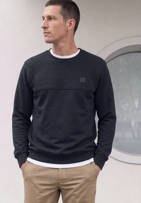 Een man draagt een zwarte trui, wit T-shirt en beige broek, met handen in de zakken.