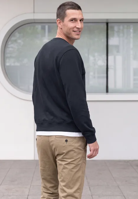 Man in zwarte trui en beige broek kijkt over zijn schouder voor een modern, licht gebouw.