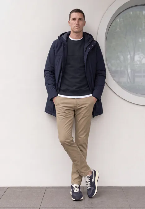 Een man leunt nonchalant tegen een witte muur met ronde ruit, in jas, trui en chino’s.