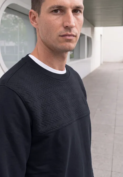Een man in een zwarte trui met geometrisch patroon staat voor een modern gebouw.