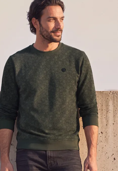 Mann mit dunkelgrünem Pullover, Bart und dunklen Haaren, steht seitlich vor einer Mauer im Freien.