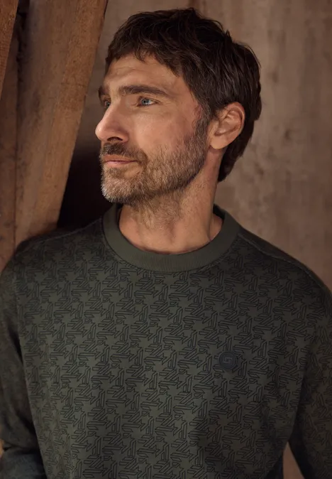 Ein bärtiger Mann in einem dunkelgrünen Pullover mit Muster blickt seitlich vor eine rustikale Wand.