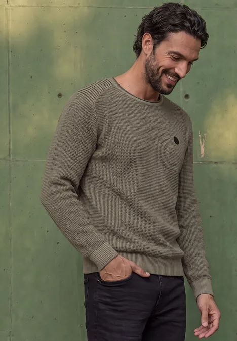 Un homme souriant porte un pull kaki et un pantalon noir devant un mur vert.