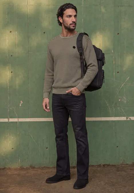 Un homme en pull vert, jean sombre et sac à dos se tient devant un mur vert.