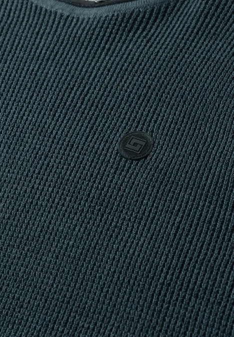 Close-up van een donkergrijze gebreide trui met V-hals en een klein logo-embleem op de borst.