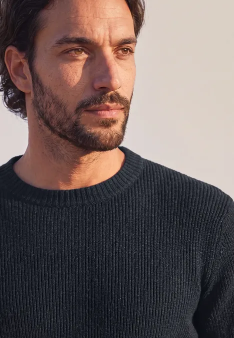 Portret van een peinzende man met donkere trui, baard en licht golvend haar.