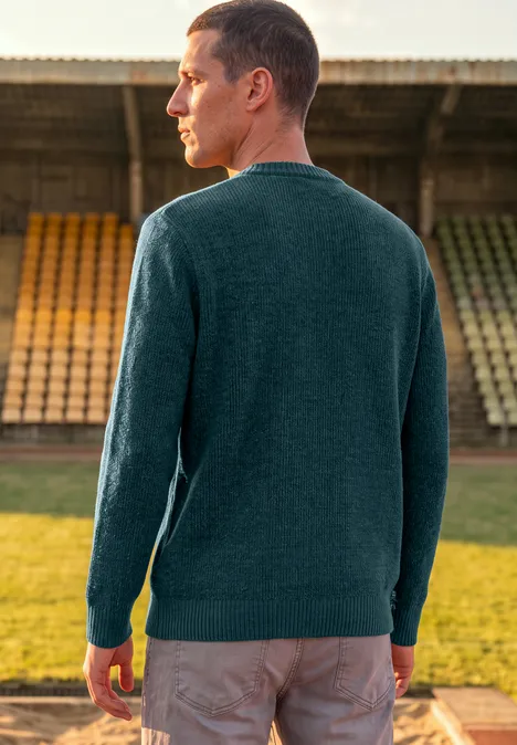 Mann im grünen Pullover steht mit dem Rücken zur Kamera vor leeren Tribünen im Freien.