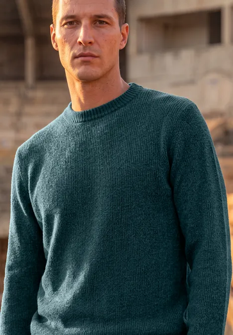 Ein Mann in einem dunkelgrünen Strickpullover steht vor einer unscharfen, urbanen Kulisse.