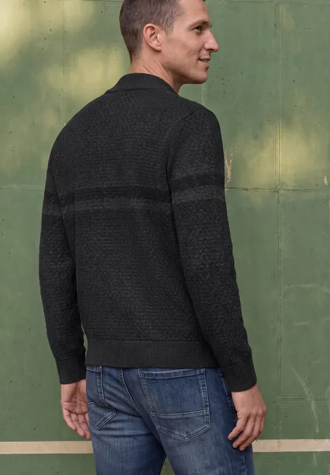Ein Mann steht mit dem Rücken zur Kamera, trägt schwarzen Pullover und blaue Jeans vor grüner Wand.