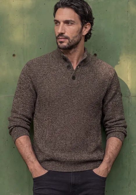 Ein Mann mit dunklen Haaren trägt einen braunen Strickpullover und steht vor einer grünen Wand.