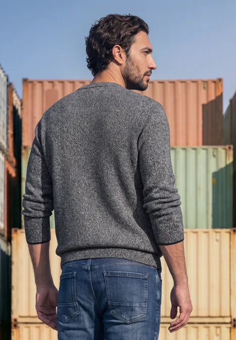 Een man in grijze trui en spijkerbroek staat met de rug naar gekleurde containers gericht.
