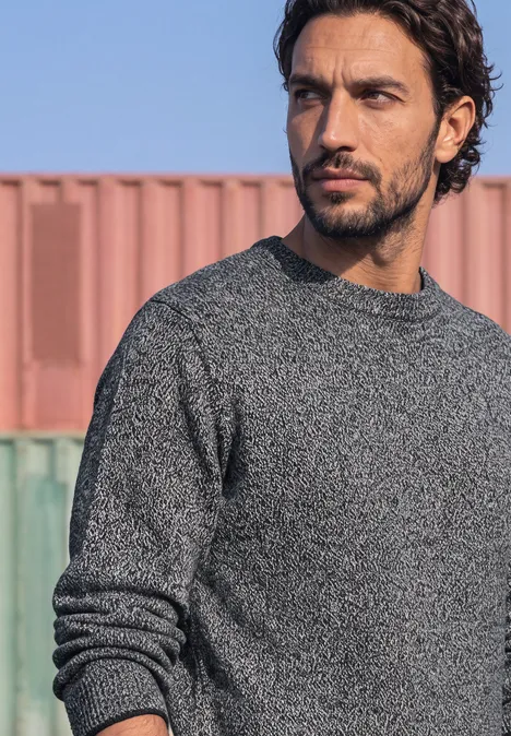 Een man met baard en donker haar draagt een grijze trui voor kleurrijke containers.