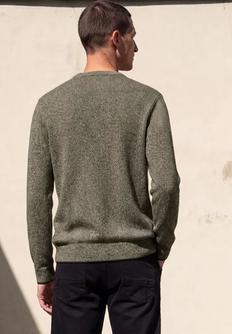 Een man staat met zijn rug naar de camera, draagt een groene trui en donkere broek voor een lichte muur.