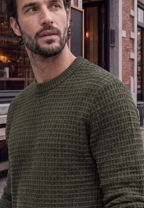 Mann mit olivgrünem, grob gestricktem Pullover vor städtischem Gebäude